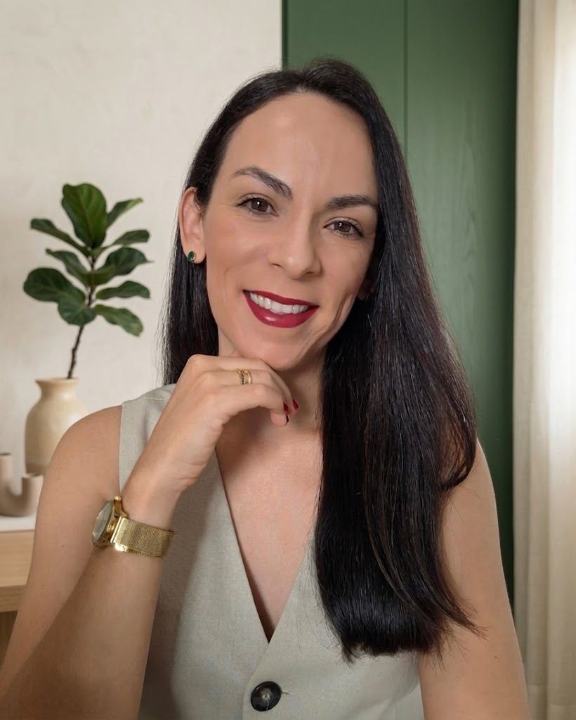 Elina Dias, nutricionista, sorrindo com a mão apoiada no queixo, de colete claro, diante de uma parede verde com uma planta ao fundo
